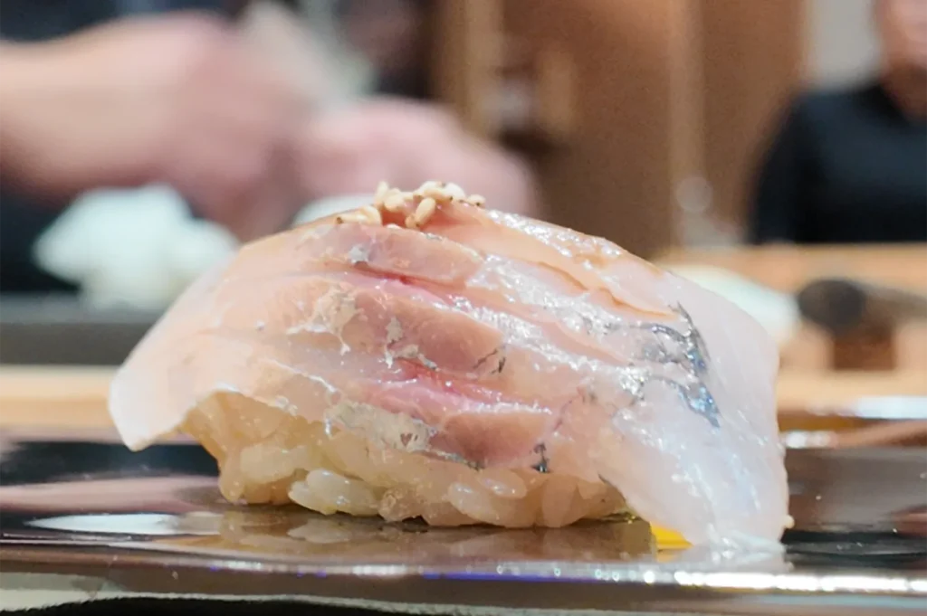 A side profile shot of a white fish nigiri (shiromi) seasoned with toasted sesame seeds on a bed of seasoned vinegared rice.