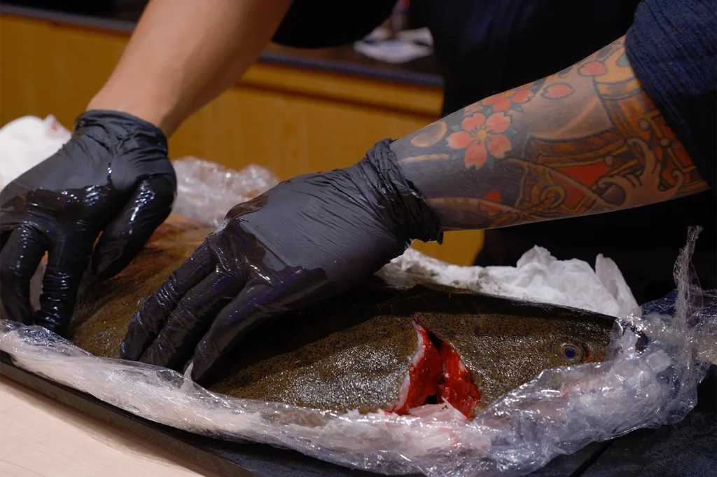 A chef with traditional floral tattoos wearing black nitrile gloves while preparing and slicing a large, fresh raw fish on a prep station.