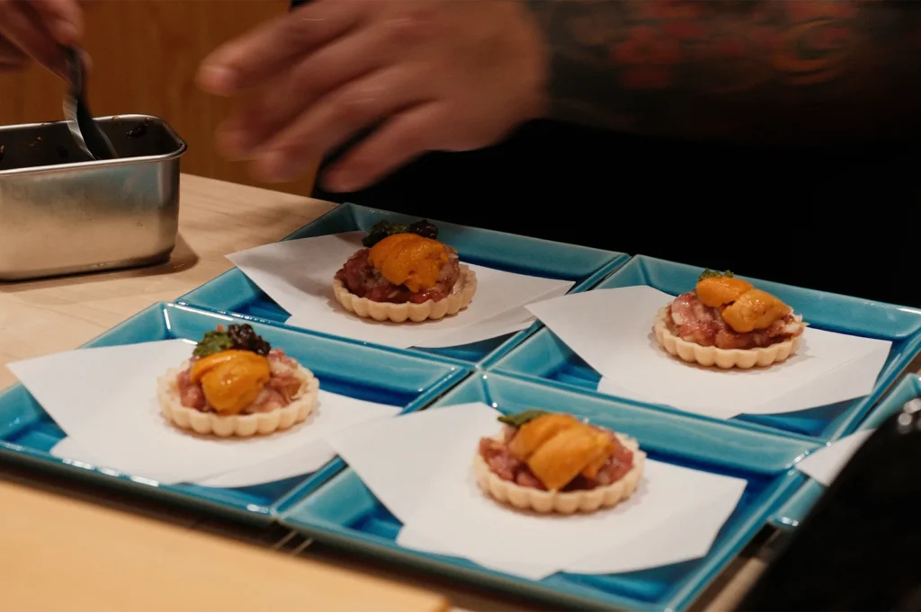Four delicate tartlets topped with wagyu beef tartare and fresh orange sea urchin (uni) being plated on turquoise square dishes.
