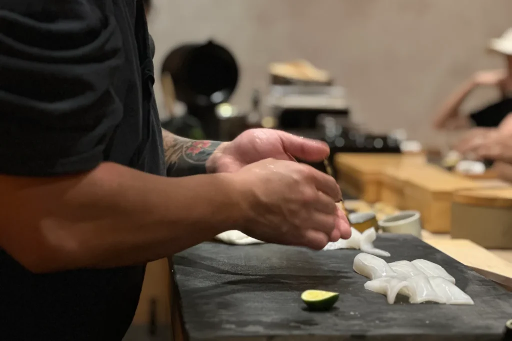 Chef Masa skillfully prepares sushi on a black cutting board, lit warmly in a cozy restaurant setting. Freshly sliced lime, glossy white fish, and high ingredient quality are visible.