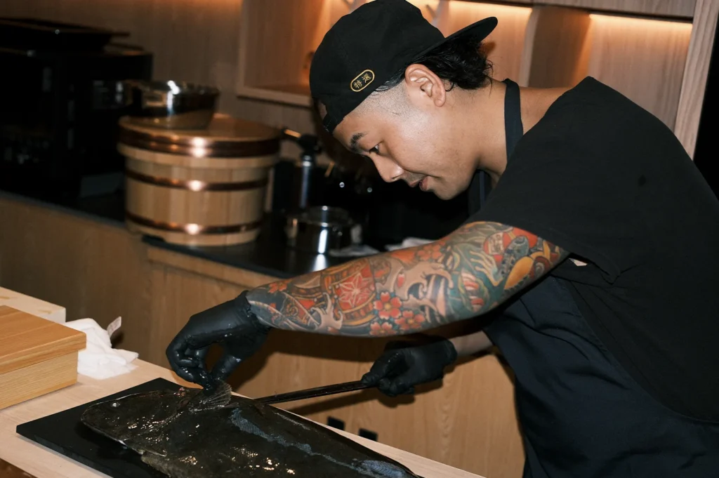 A professional sushi chef with detailed arm tattoos skillfully prepping a whole fish on a cutting board.