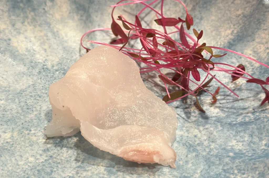 A thin, translucent slice of premium white fish sashimi garnished with delicate red microgreens on a textured light blue plate.