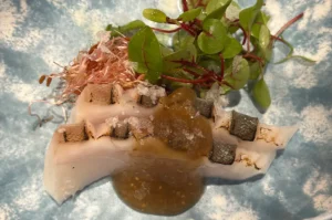 Gourmet seared white fish sashimi topped with savory miso sauce, microgreens, and radish sprouts on a blue ceramic plate.