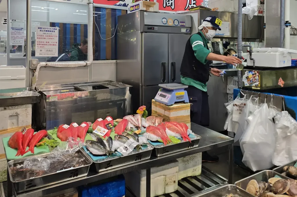 Interior of a Japanese fish market featuring whole red snapper (tai) and mackerel on ice displays with a fishmonger in the background.