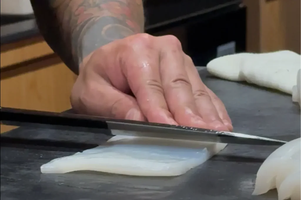 A chef with a forearm tattoo carefully scoring or slicing a piece of translucent white squid (ika) on a dark cutting board.