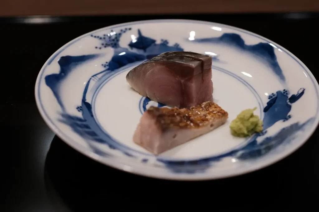 Two thick slices of seared bonito (katsuo no tataki) served with a dollop of fresh wasabi on a blue and white patterned plate.