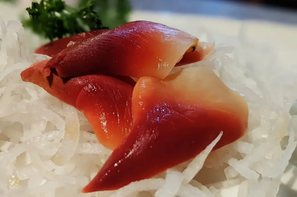 Close-up of sliced red and white clam sashimi on a bed of thinly sliced daikon radish. Fresh parsley garnish in the background adds a touch of green.