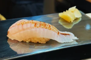 A piece of lightly grilled sushi with white fish on rice, set on a shiny metallic plate. In the background, there are slices of pickled ginger.