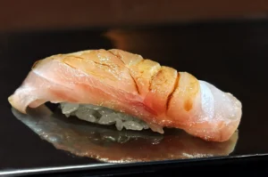 A piece of sushi featuring a slice of pink and white fish atop neatly formed white rice, on a reflective black surface, conveying freshness and elegance.