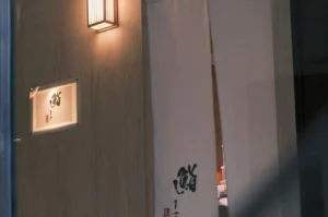 Japanese restaurant entrance with beige curtains featuring calligraphy, a glowing wall lamp, and a sign, creating a serene, welcoming ambiance.