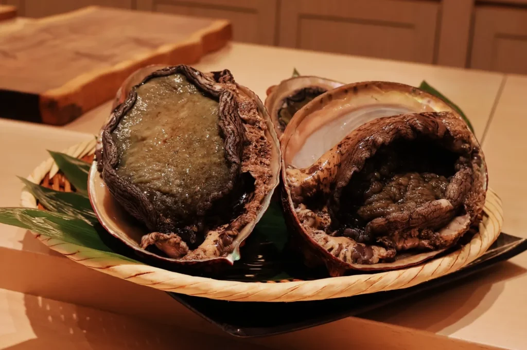 Basket with two large abalone, shells slightly open, on a wooden table. The texture appears rough; green leaves underneath add freshness.
