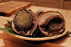 Basket with two large abalone, shells slightly open, on a wooden table. The texture appears rough; green leaves underneath add freshness.