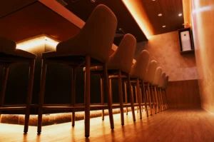 Row of tall chairs in a modern bar setting, dimly lit with warm tones. Soft lighting creates a cozy, inviting atmosphere.