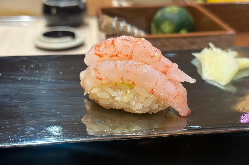 A single piece of Ama Ebi (sweet shrimp) nigiri sushi garnished with lime zest, served on a black lacquered plate with pickled ginger.