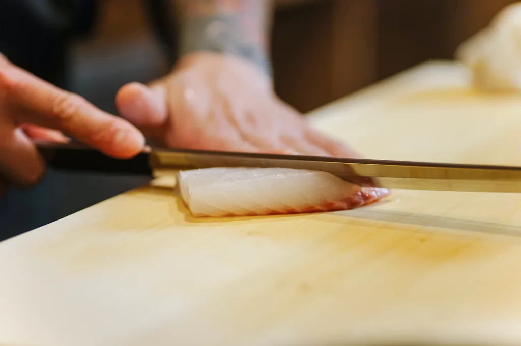 Close-up of a chef's hands using a traditional Japanese knife to precisely slice a fresh piece of white fish on a wooden cutting board for sashimi.