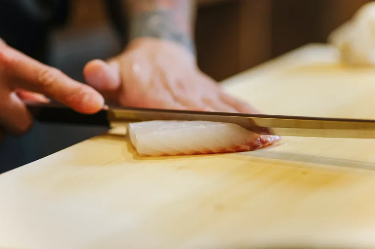 Close-up of a chef's hands using a traditional Japanese knife to precisely slice a fresh piece of white fish on a wooden cutting board for sashimi.