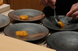 A professional Japanese chef carefully plating fresh orange sea urchin (uni) onto dark textured ceramic plates using a spoon.