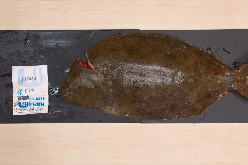 A whole fresh Japanese Hirame (Olive Flounder) lying on a black prep board next to a Japanese quality certification label.