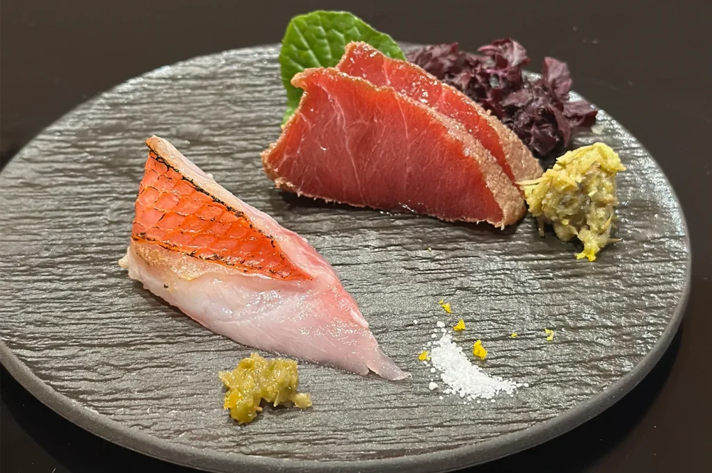 A gourmet sashimi platter featuring a piece of seared Kinmedai (Golden Eye Snapper) and sliced bluefin tuna, served with fresh wasabi and sea salt on a textured stone plate.