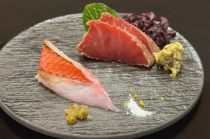 A gourmet sashimi platter featuring a piece of seared Kinmedai (Golden Eye Snapper) and sliced bluefin tuna, served with fresh wasabi and sea salt on a textured stone plate.