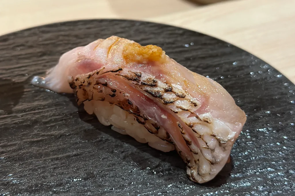 A piece of premium Nodoguro (Blackthroat Seaperch) nigiri sushi, lightly seared (aburi) to render the fat, served on a textured black plate.