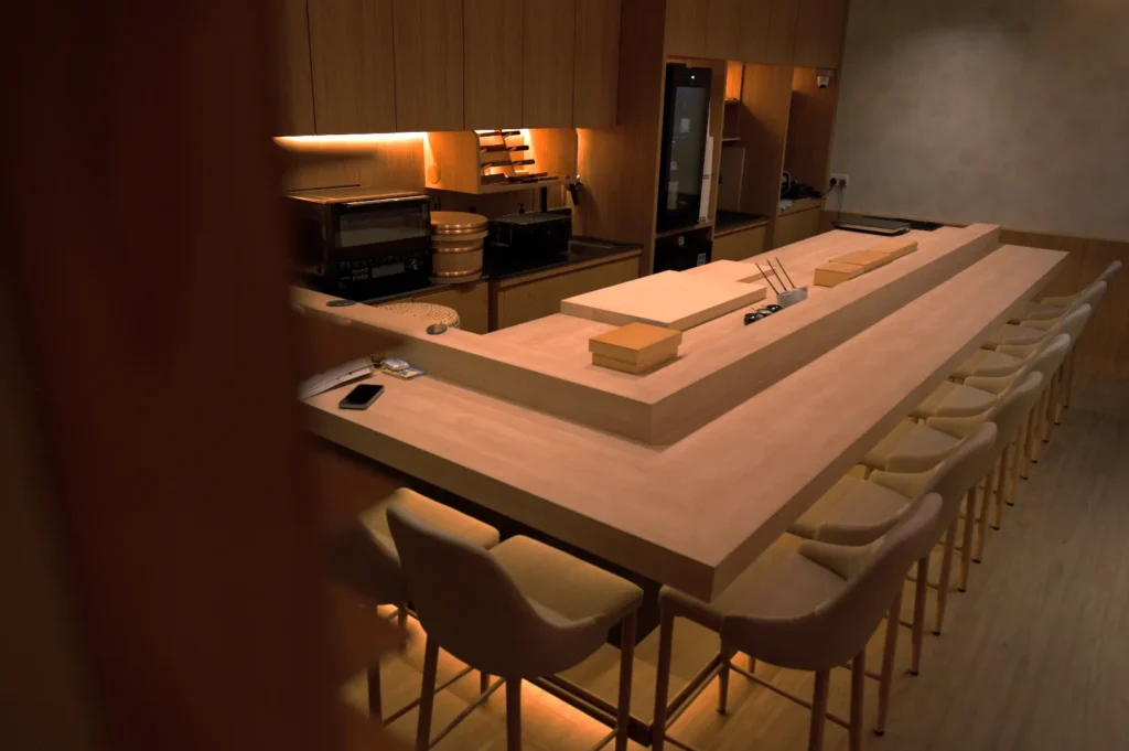 Dimly lit wooden kitchen with a long island counter, high-back chairs, and warm lighting. Minimalistic design evokes a cozy, intimate atmosphere.