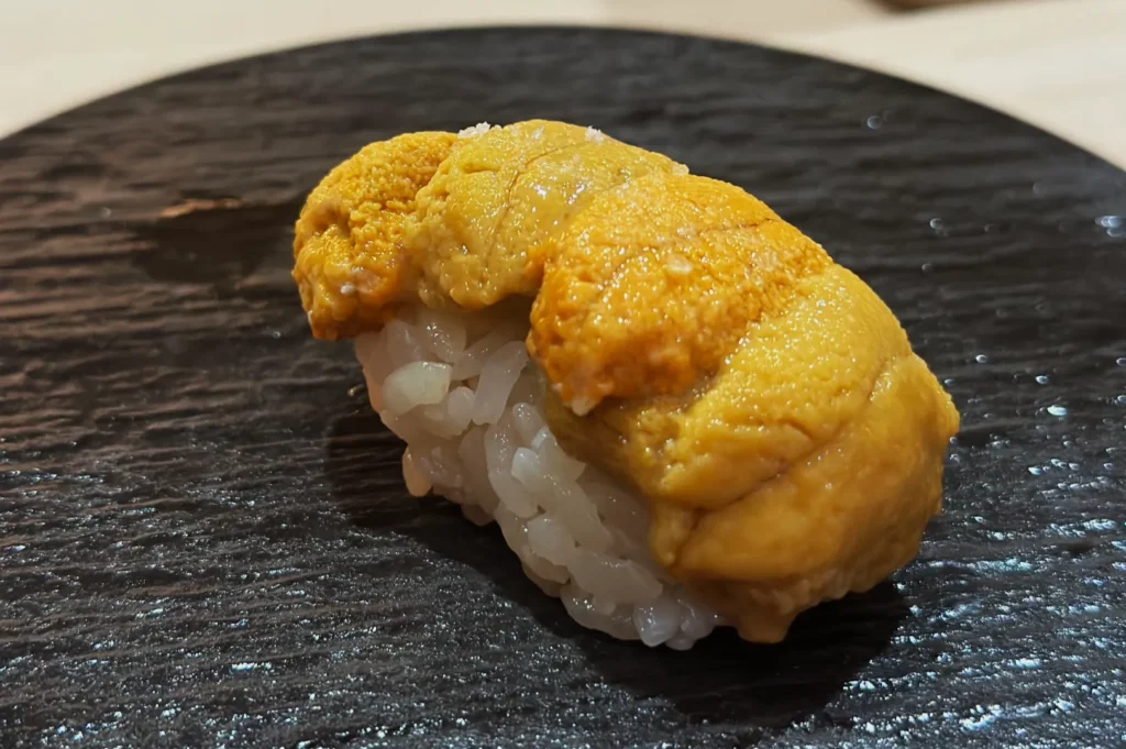 Close-up of a piece of uni sushi on a textured black plate. The golden sea urchin rests atop a small mound of white rice, creating a gourmet presentation.