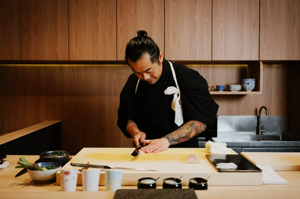 Chef in black attire skillfully slices fish on a wooden counter in a modern kitchen. The setting is minimalist, focused, and elegant.