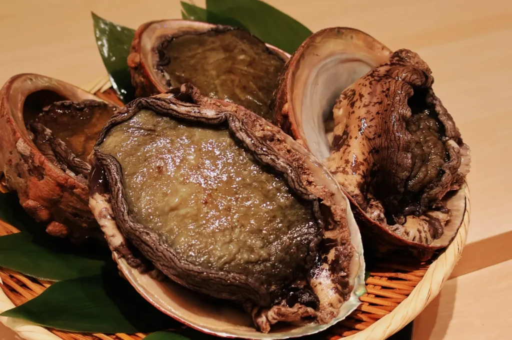 A basket of three fresh, open abalone shells resting on green leaves. The image highlights their glossy, textured flesh and rugged shells, conveying a sense of freshness and raw natural beauty.