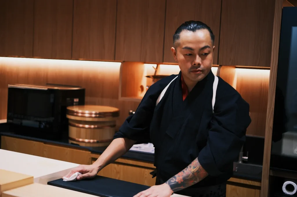 A chef in a black uniform cleans a counter in a modern kitchen, focused and attentive. Warm lighting highlights the wooden cabinets and cooking equipment.