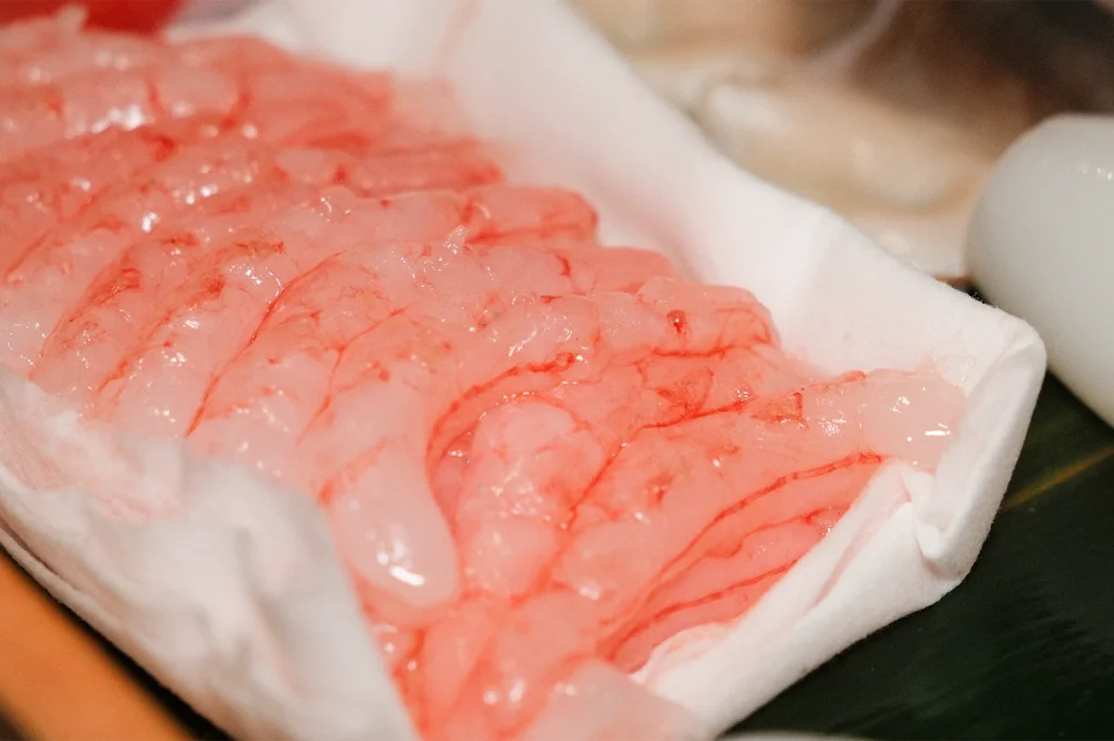 A close-up view of fresh, translucent raw pink sweet shrimp (Amaebi) arranged neatly on a white paper-lined tray.