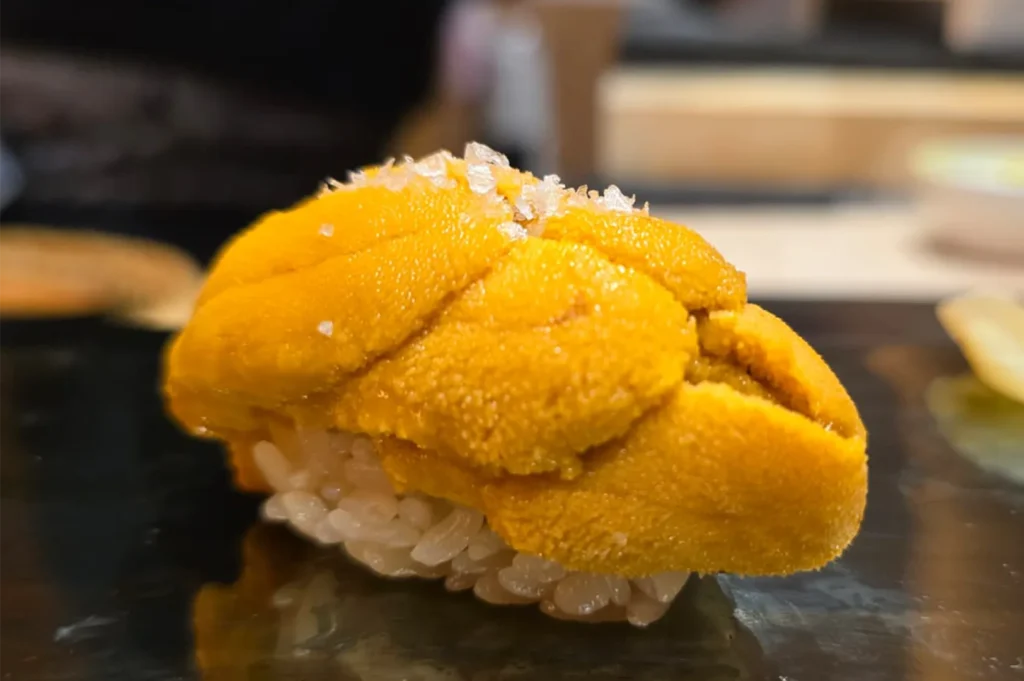 A close-up shot of premium Uni (sea urchin) nigiri sushi topped with delicate flakes of sea salt, showcasing the fresh, creamy texture of the roe.