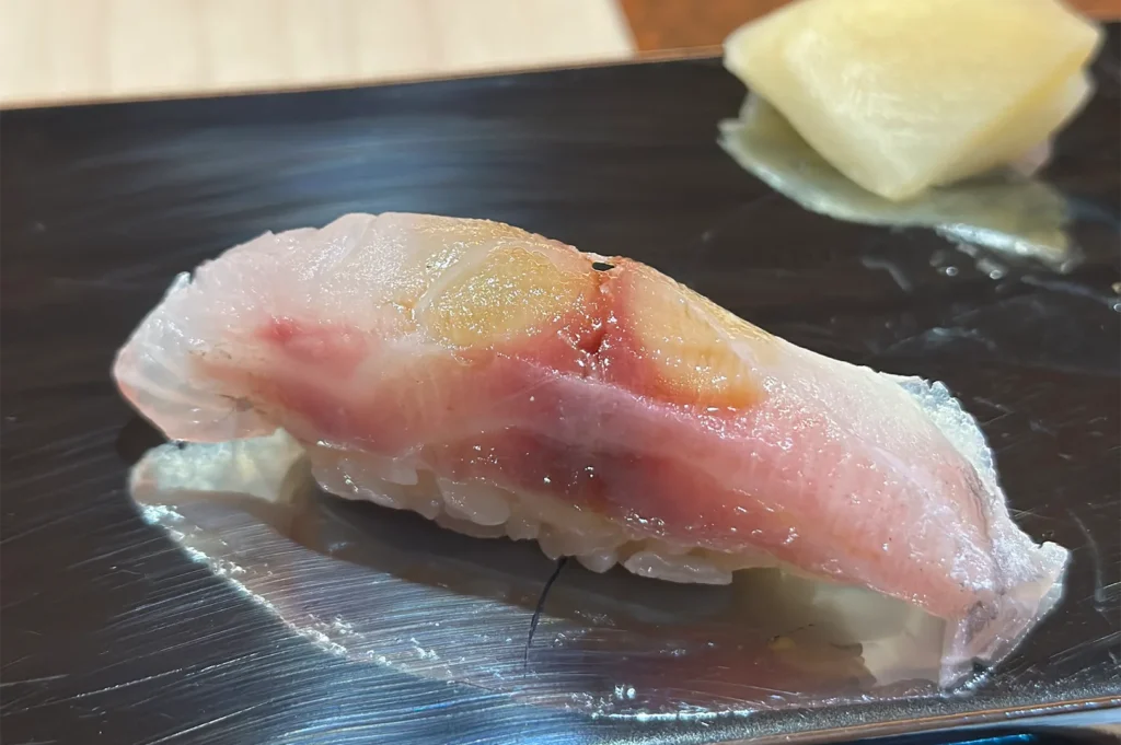 A piece of Shima Aji (striped jack) nigiri sushi with a pinkish hue, served on a sleek black lacquer plate with pickled ginger.