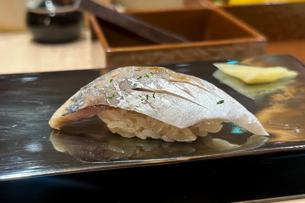 A close-up shot of Shime Saba (cured mackerel) nigiri sushi with silver skin, served on a black lacquer board with pickled ginger.