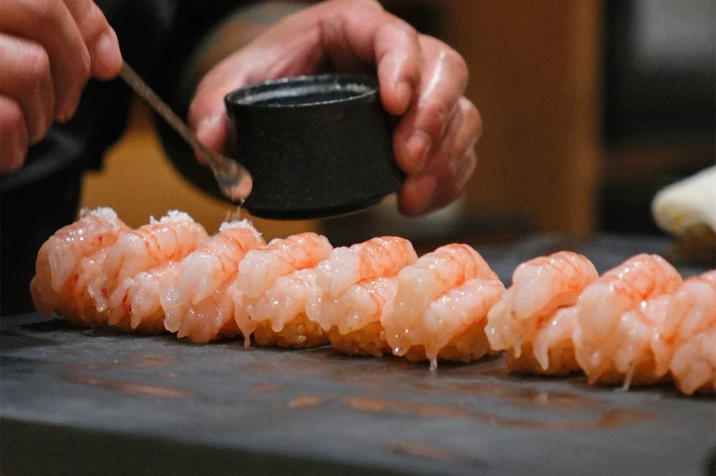 Close-up of a sushi chef using a small spoon to carefully apply salt or seasoning to a row of fresh shrimp nigiri.
