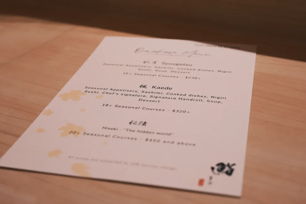 Elegant menu on a wooden table lists omakase courses with prices; delicate, autumn-themed foliage accents create a refined, seasonal feel.