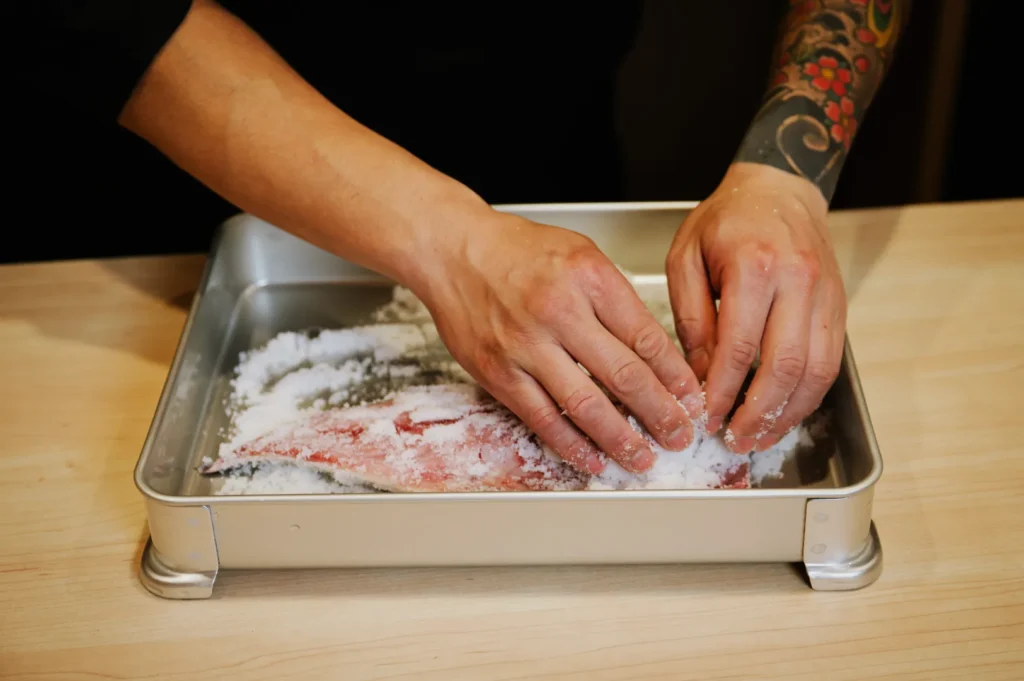 Hands sprinkle salt over a fish fillet in a metal baking tray on a wooden countertop. The scene conveys a sense of focus and culinary preparation.