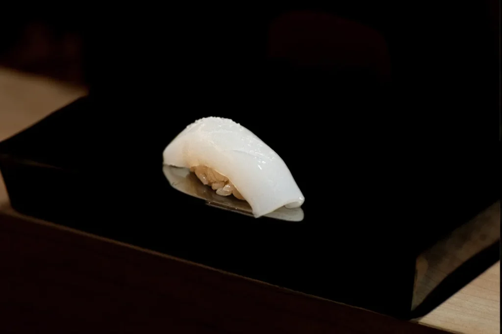 A single piece of delicate white sushi, placed on a reflective black surface, showcases rice topped with a tender slice of fish. The setting is minimalistic and elegant.