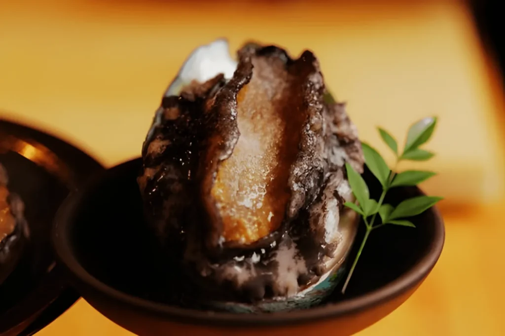 A close-up of a fresh abalone in its shell, served in a black bowl. The glossy, textured surface is highlighted with a small green garnish. The setting is elegant and warm.