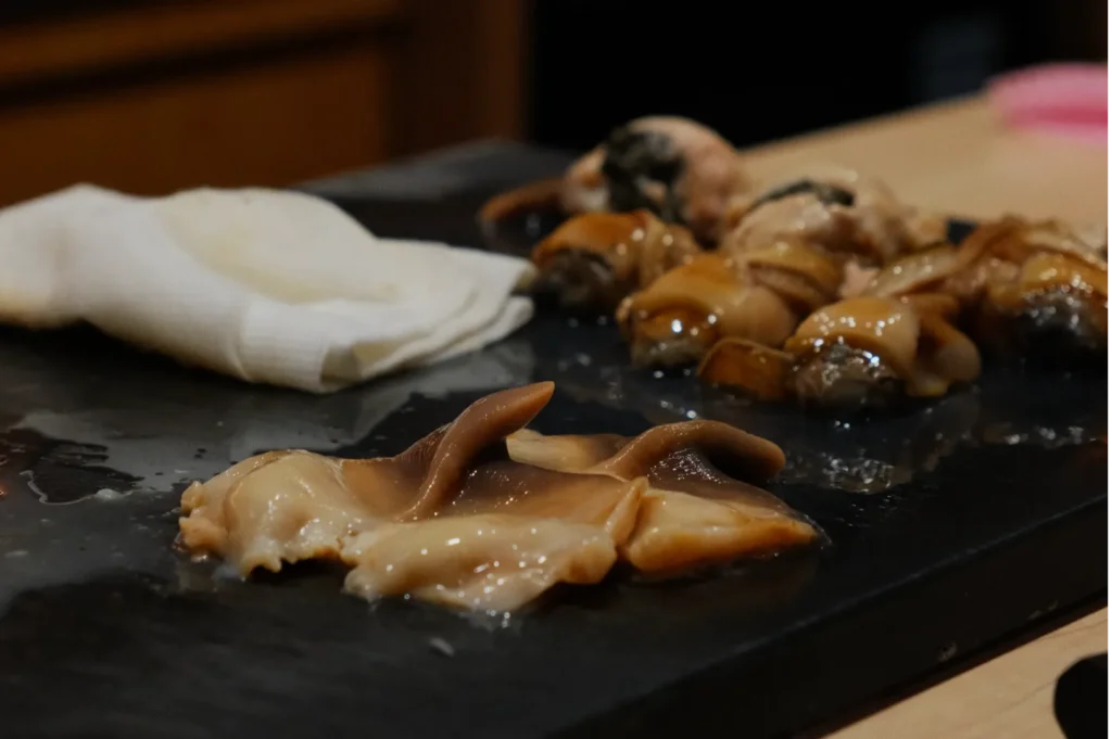 Close-up of raw seafood on a dark slate board, featuring sliced shellfish and several conch shells. A folded white napkin lies beside them.