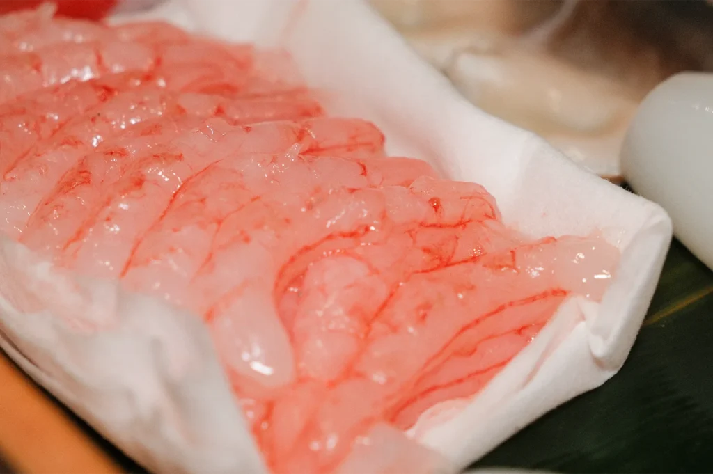 A pile of fresh, translucent raw peeled sweet shrimps resting on a white refrigerated tray, ready for sushi preparation.