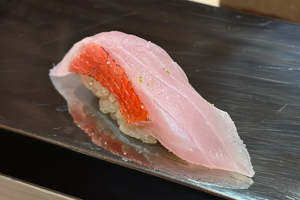 Kinmedai (Golden Eye Snapper) nigiri sushi featuring its signature bright red skin and delicate pink flesh, garnished with citrus zest.