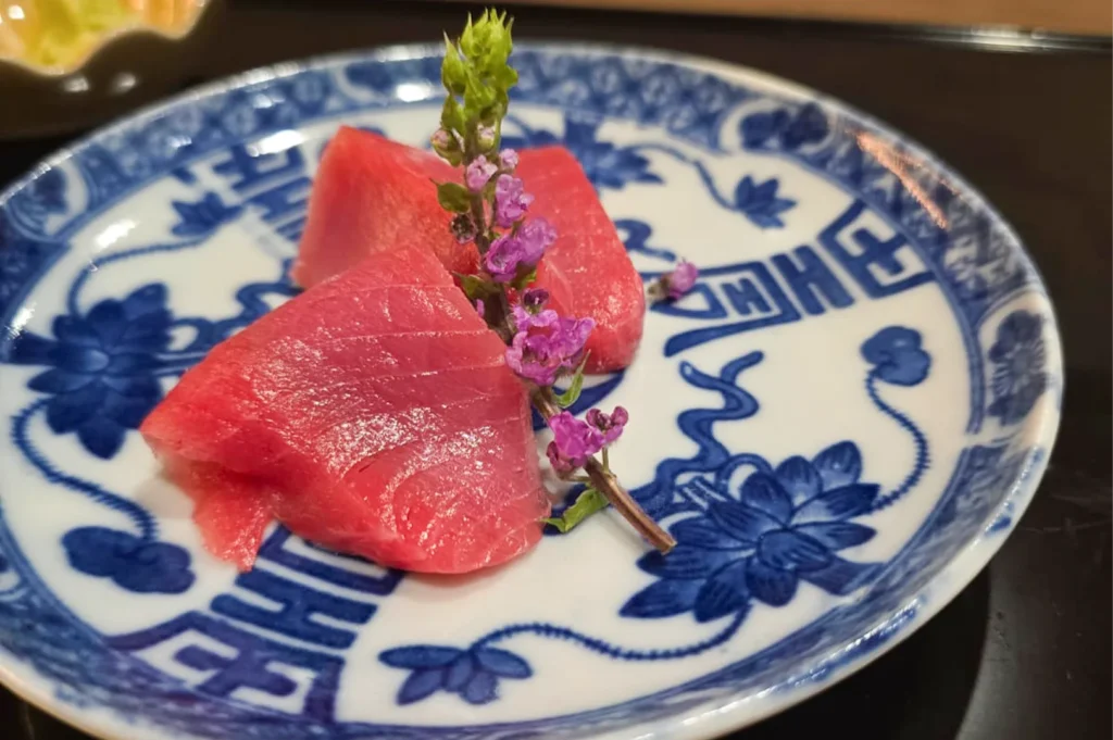 Two thick slices of fresh red maguro tuna sashimi served on a traditional blue and white patterned Japanese plate, garnished with a purple shiso flower.