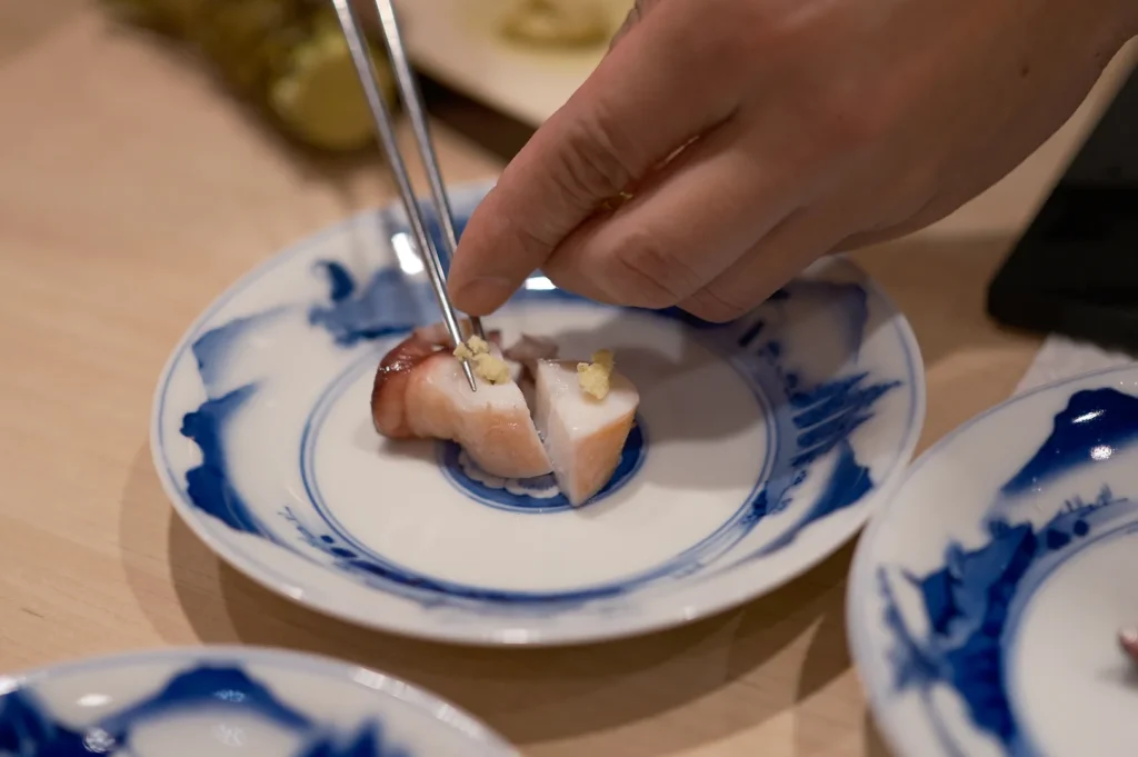 A sushi chef using silver chopsticks to precisely place fresh wasabi onto sliced boiled octopus (tako) on a blue patterned plate.