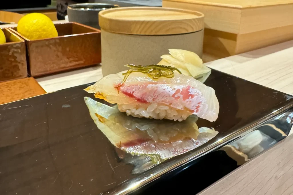 Sea bream (Tai) nigiri sushi topped with a sliver of kelp (kombu) and a light glaze, presented on a reflective black serving board.