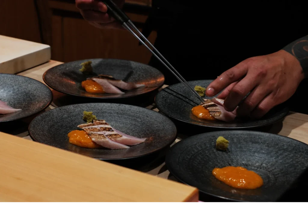 Chef's hands expertly place thin slices of seared fish on dark plates, each accompanied by a dollop of orange sauce and green wasabi, conveying precision and elegance.