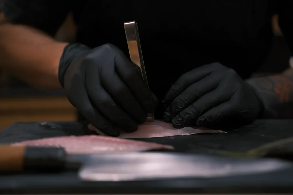 A person wearing black gloves uses tweezers to prepare raw fish on a cutting board. A knife is visible in the foreground. The scene is focused and precise.