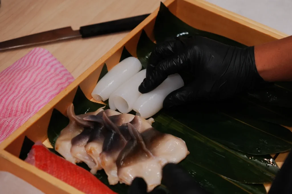 A person wearing black gloves arranges white fish fillets and squid rolls on banana leaves in a wooden tray. A knife and pink striped cloth are nearby.