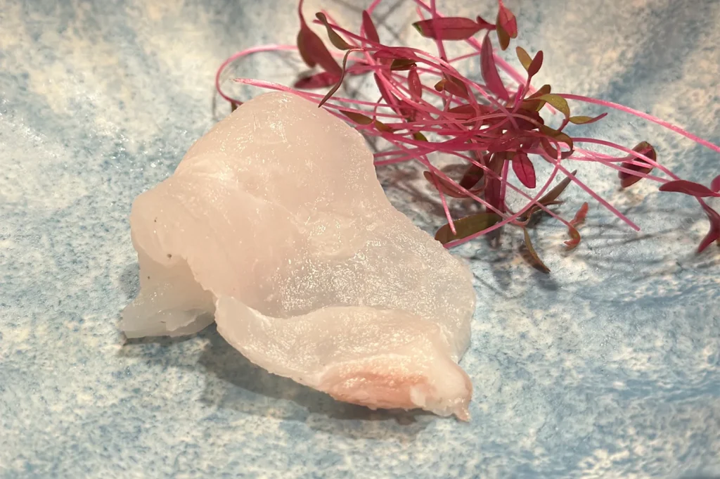 Thinly sliced Hirame (flounder) sashimi garnished with vibrant red micro-greens on a speckled light blue plate.