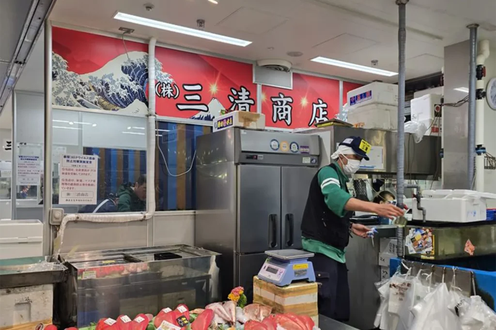 A Japanese fishmonger wearing a mask and cap working at a seafood stall with a large "Great Wave" and Mount Fuji backdrop.